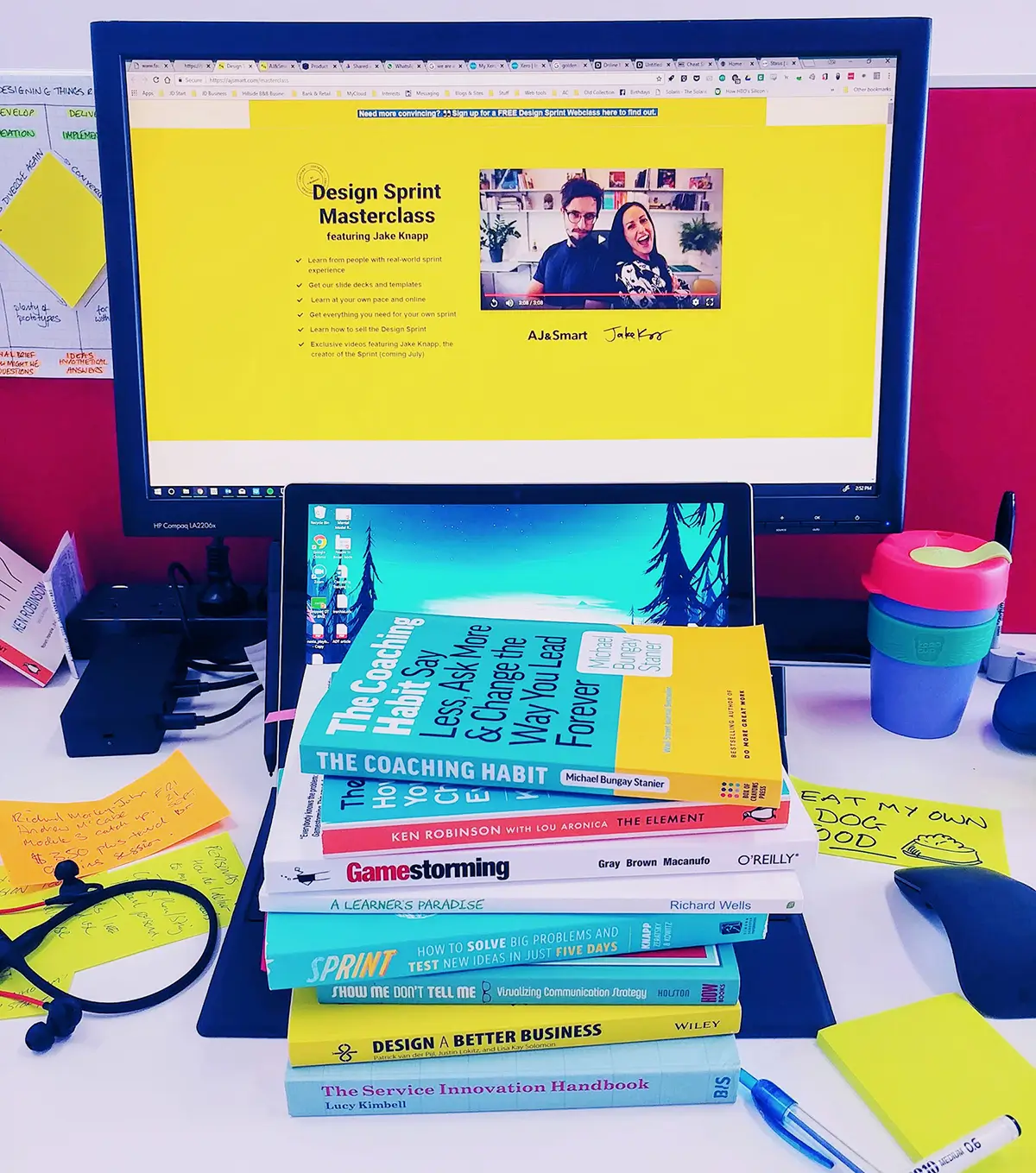 A computer screen shows a design sprint website on the screen. A pile of books on this subject are stacked on the desk in front of the screen.