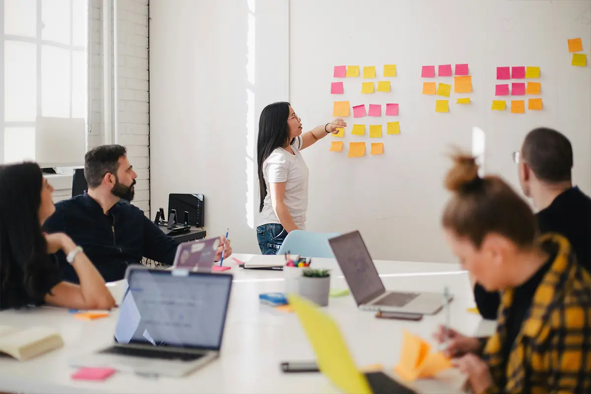 A person stands at the front of a group of people around a table, pointing to post-it notes that have been stuck to a wall